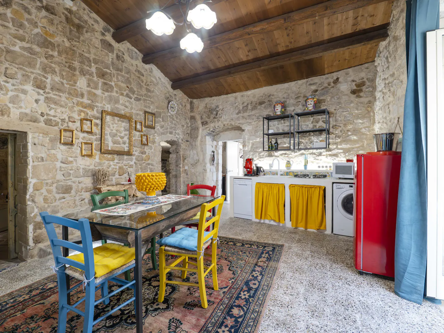 Rustic kitchen with stone walls, wooden ceiling, glass dining table, colorful chairs, and a bright red refrigerator.