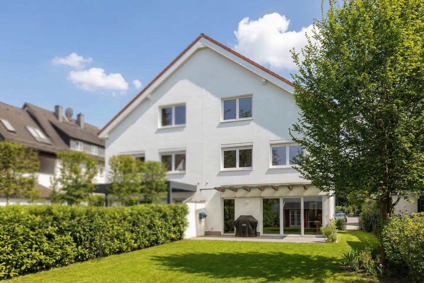 Two-story white house with a red roof, green lawn, and a patio with a grill on a sunny day.