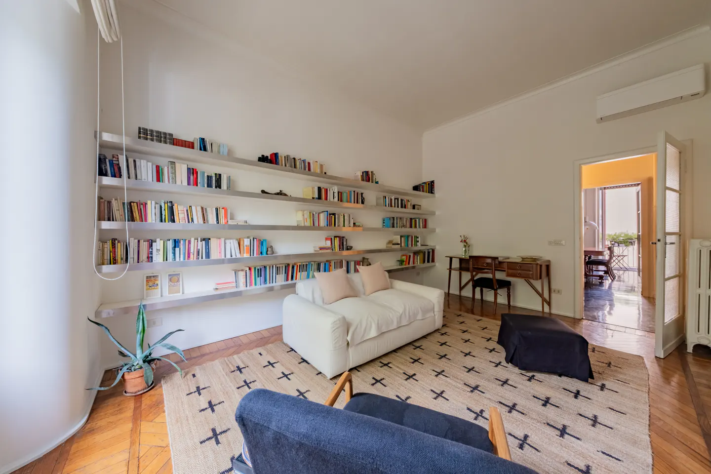 Living room with white sofa, patterned rug, wall-to-wall bookshelves, a desk, and a view into an adjacent room.