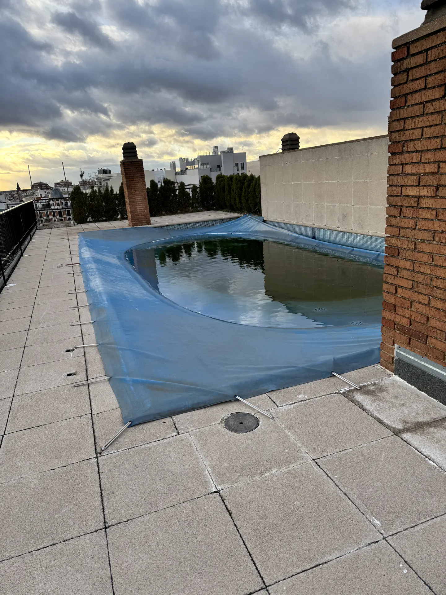 Rooftop pool covered with a blue tarp on a cloudy day. The pool is surrounded by gray tiles and a brick wall. City buildings are in the background.