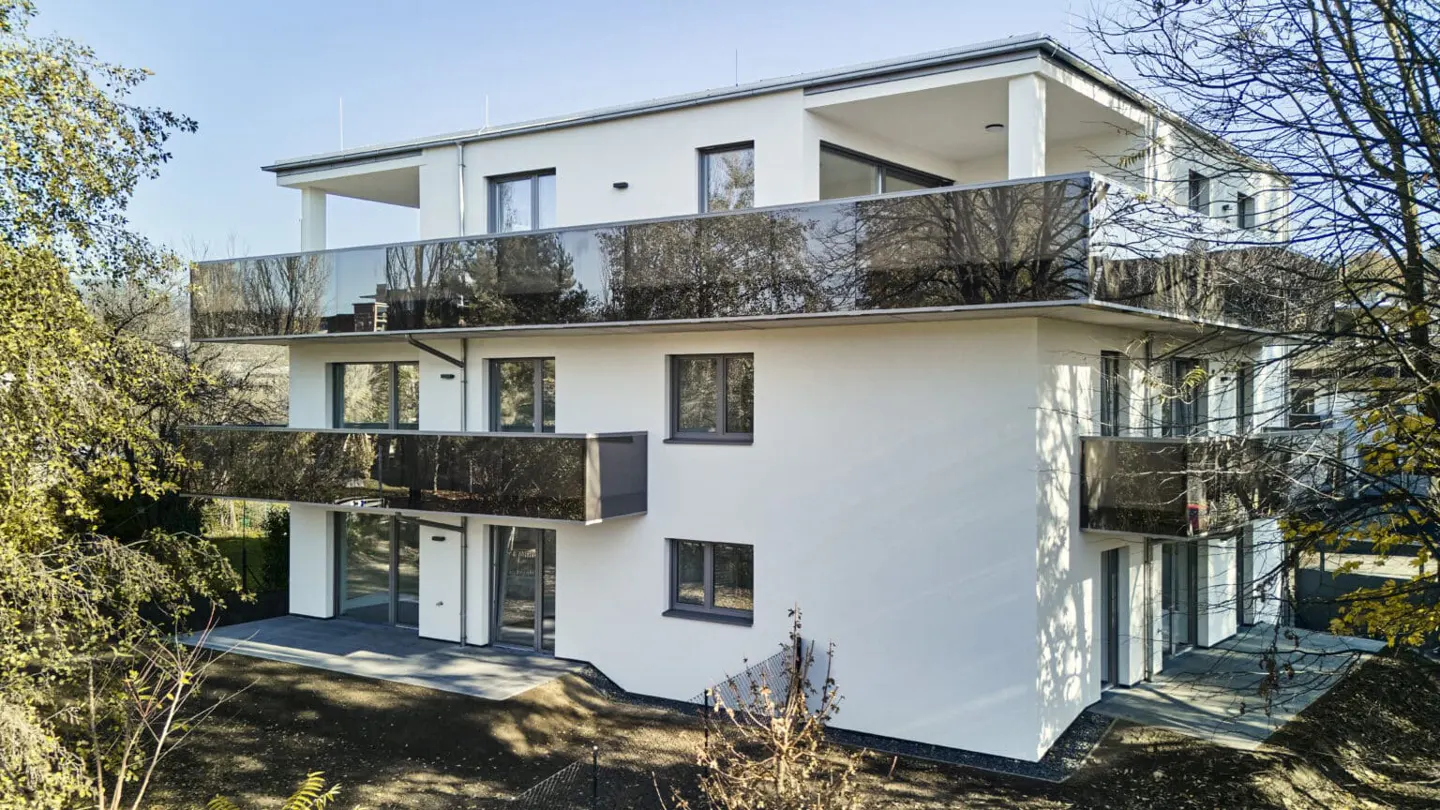 Modern white three-story apartment building with dark glass balconies and large windows, surrounded by trees.