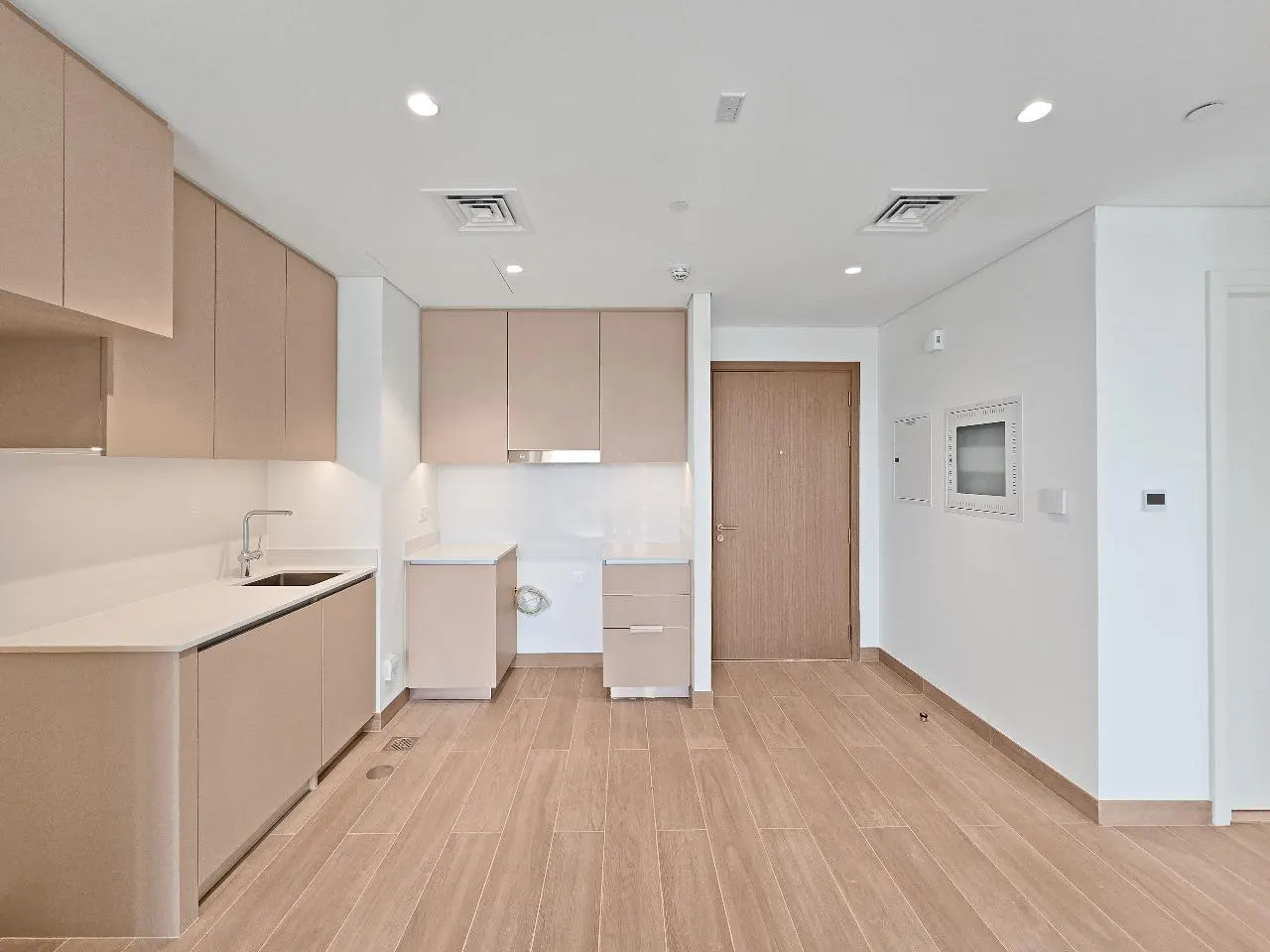 A bright, modern kitchen with light wood cabinets, white countertops, and wood-look tile flooring. A closed wooden door is on the right.