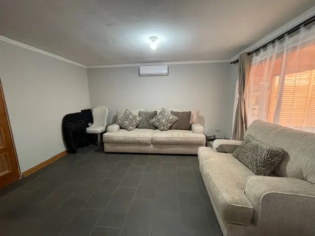 Living room with gray tile floor, beige sofas with patterned pillows, white chair, and sheer curtains.