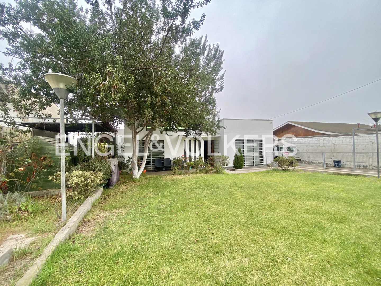 A white modern house with a large green lawn, a mature tree, and outdoor lighting under a cloudy sky.