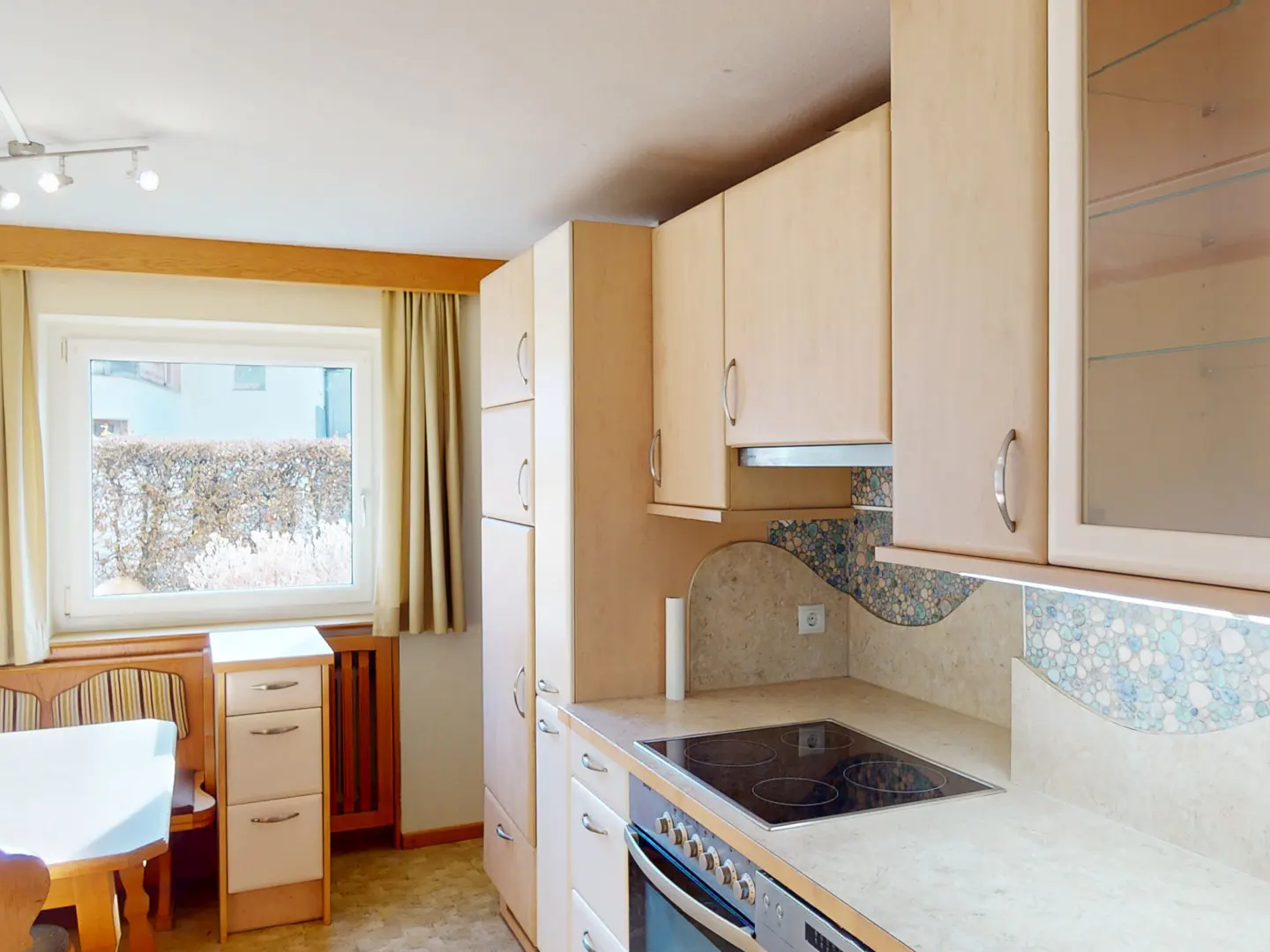 A bright kitchen with light wood cabinets, a black stovetop, and a window with beige curtains.