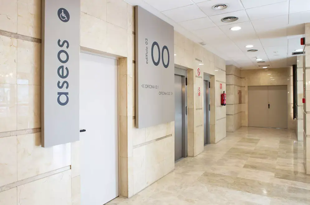 Hallway with marble floors and walls. Signs indicate restrooms and office locations. Elevators and a fire extinguisher are visible.