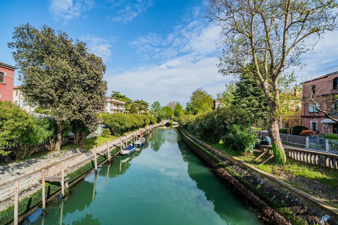 A canal with turquoise water runs between buildings and trees under a blue sky with clouds. Boats are docked along the canal's edge.