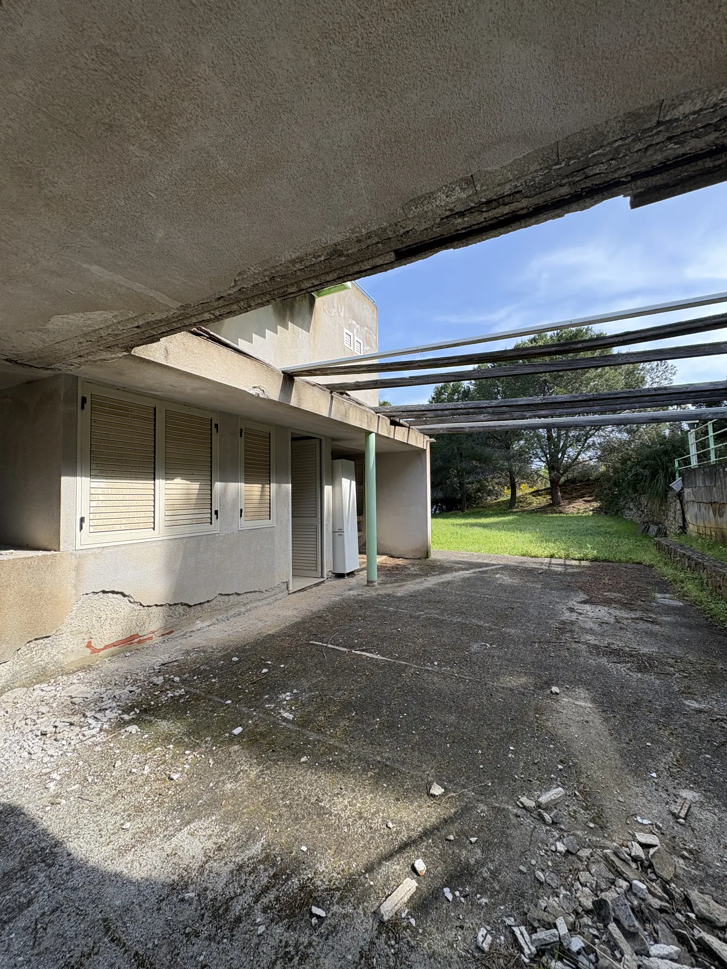 Exterior view of a weathered, concrete building with closed shuttered windows and a covered patio area. A grassy lawn and trees are visible in the background.
