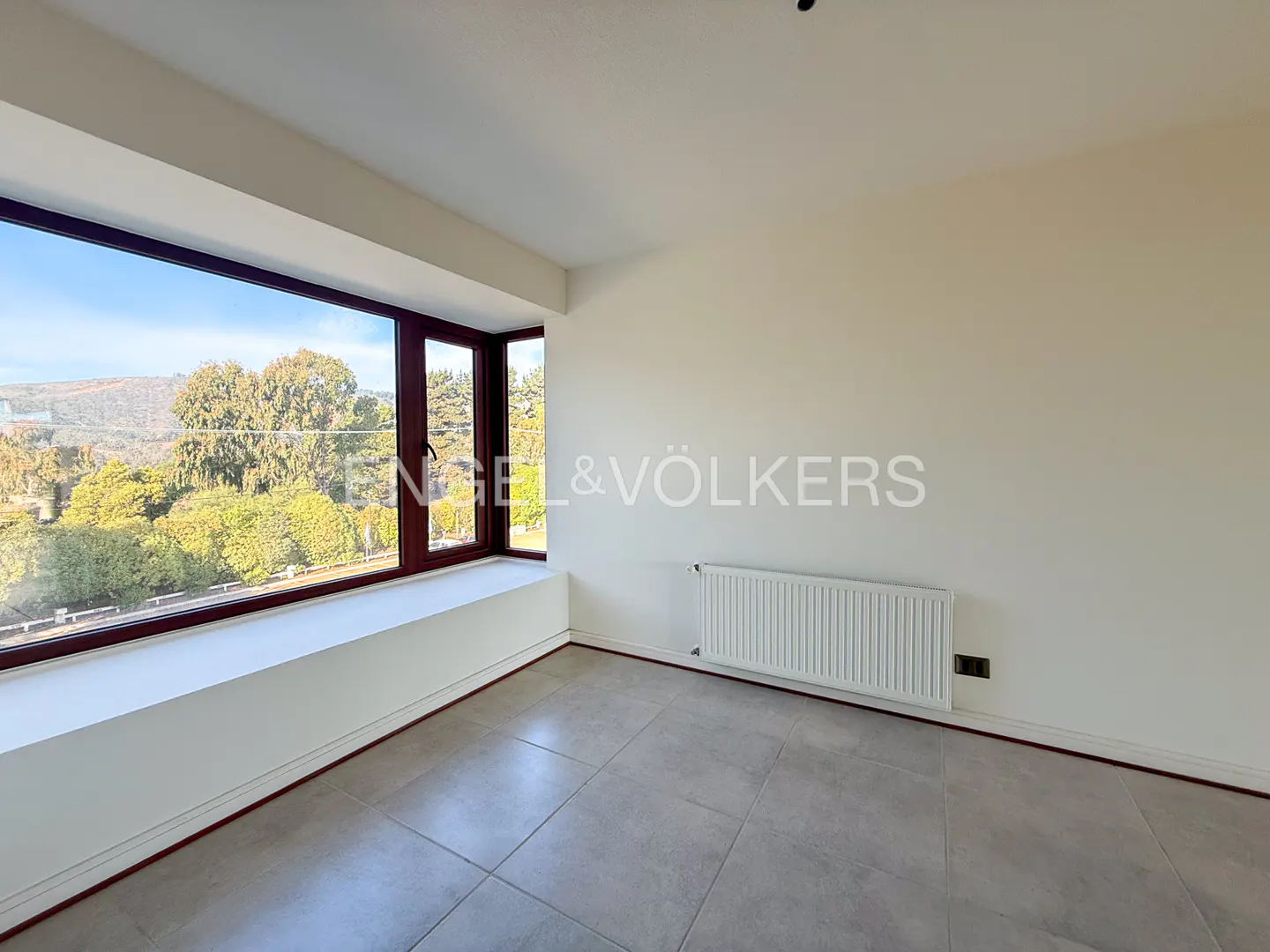 An empty room with gray tile floors, white walls, and a large window with a view of trees. A white radiator is on the wall.