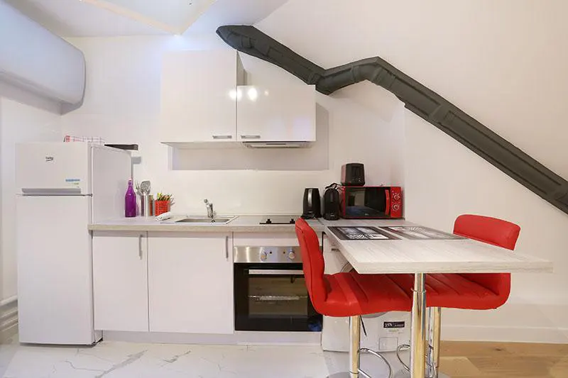 Small white kitchen with a refrigerator, oven, sink, and a breakfast bar featuring two bright red stools.