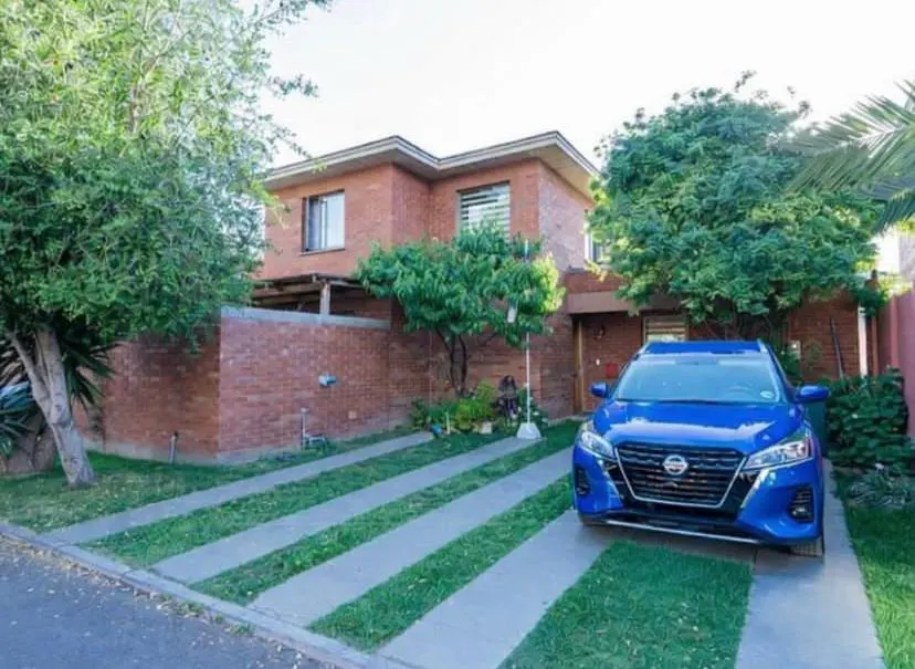 Two-story brick house with a blue car parked in the driveway. Green trees surround the house.