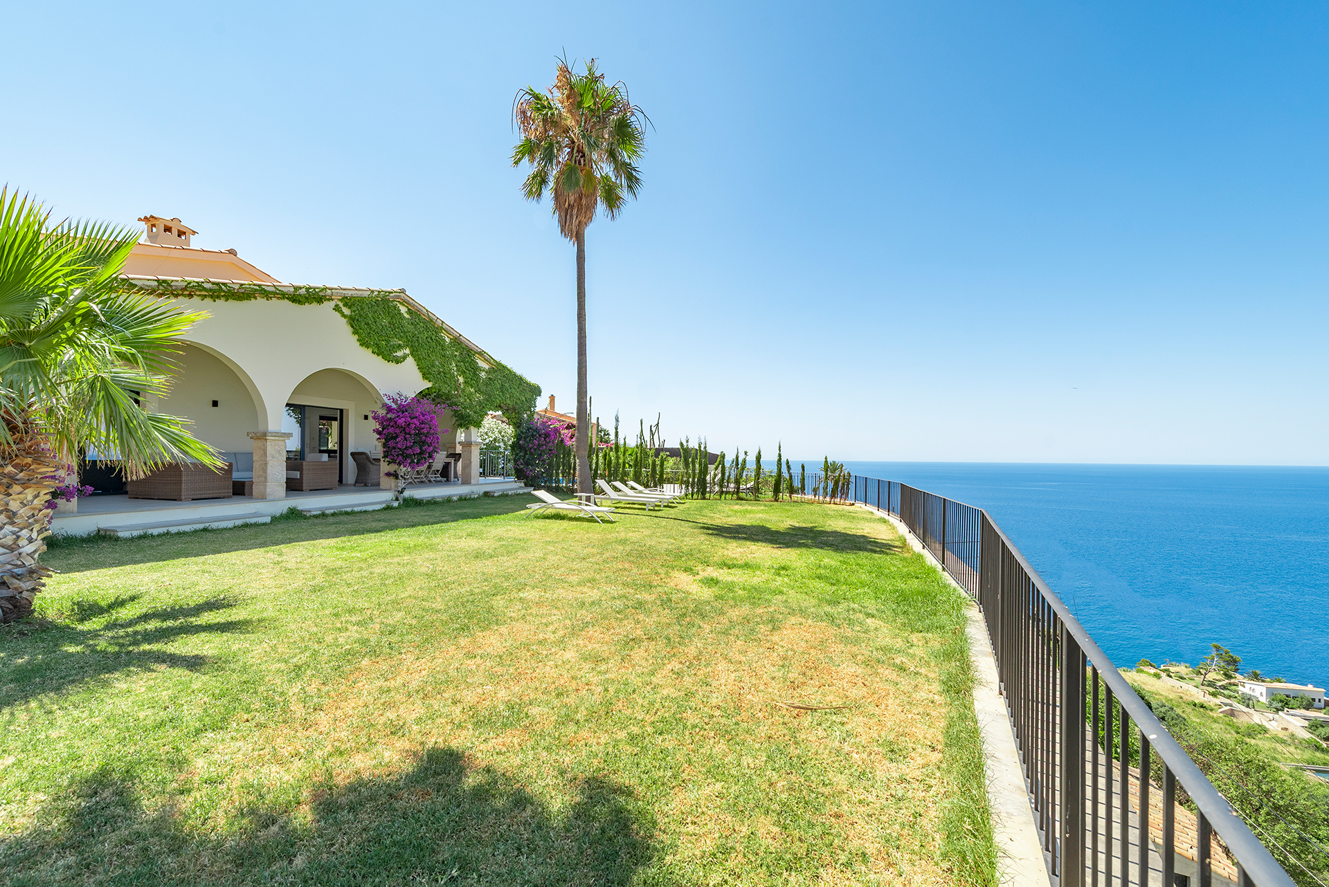 A villa with a vine-covered arched patio, palm trees, and a grassy lawn overlooking the blue ocean on a sunny day.