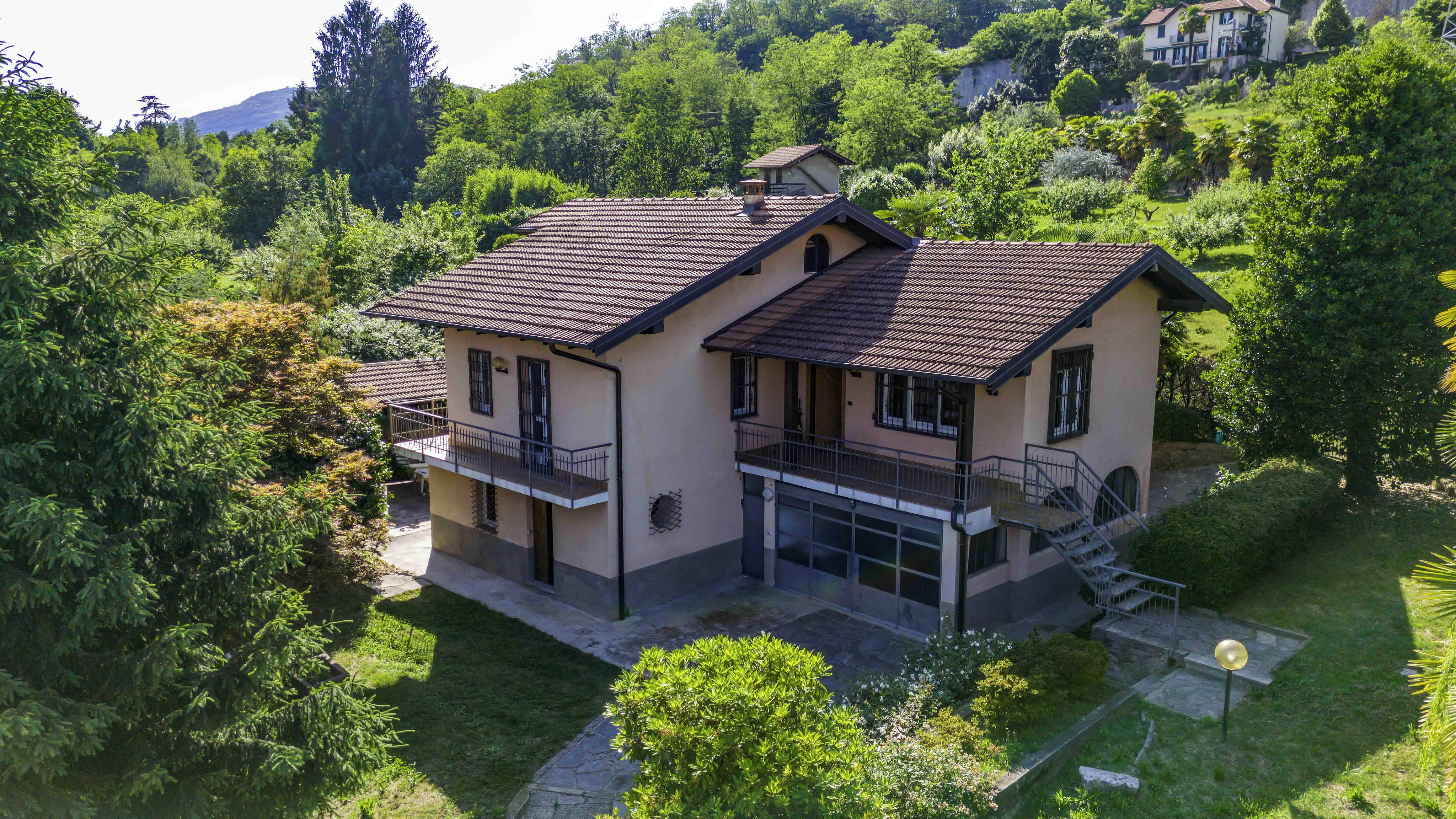 A two-story pink house with a brown tiled roof, balconies, and metal stairs, surrounded by lush green trees and gardens.