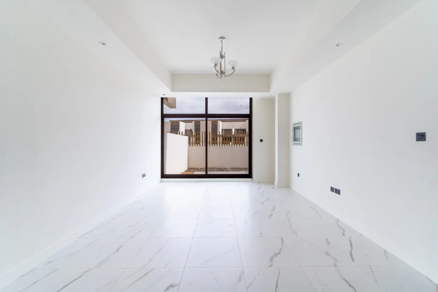 Bright, empty room with white walls and marble tile floor. A large sliding glass door leads to a fenced patio. A chandelier hangs from the ceiling.