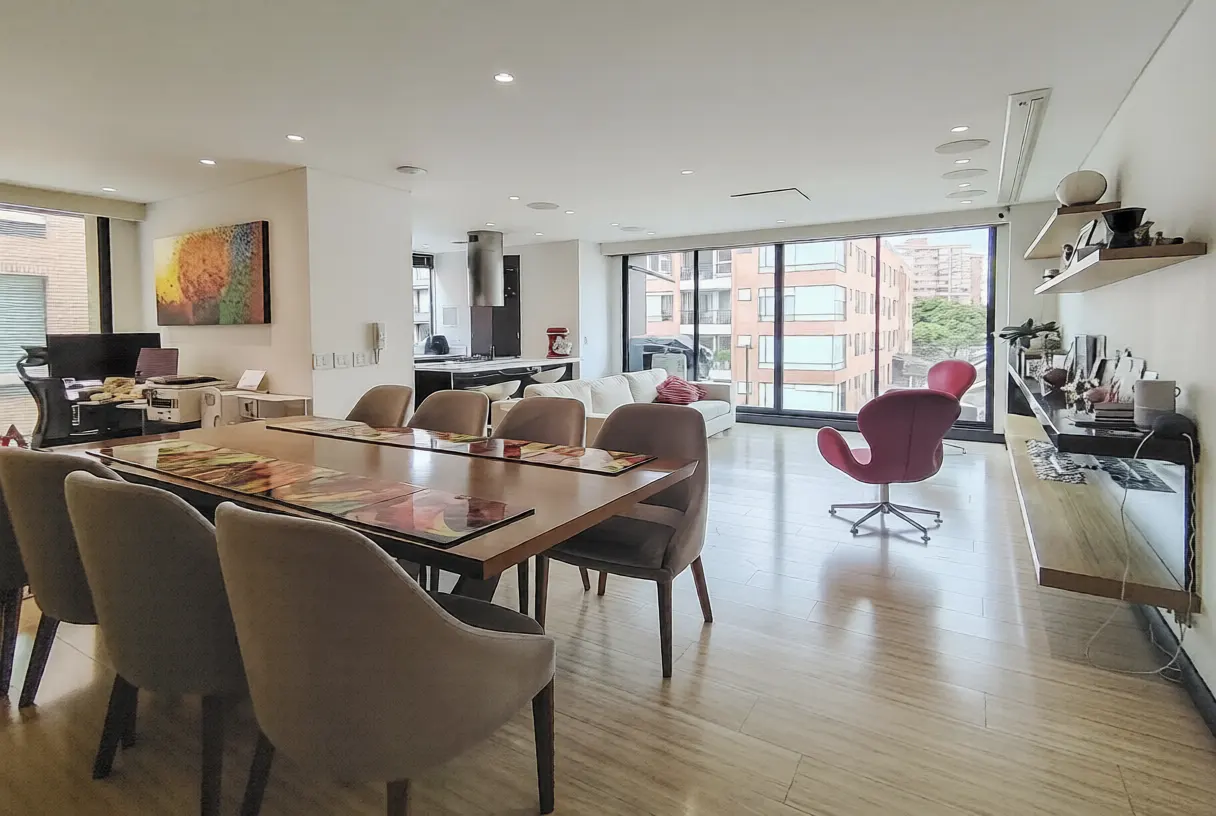 Open-concept living space with a dining table, chairs, kitchen, sofa, and a pink swan chair. Large windows offer city views.