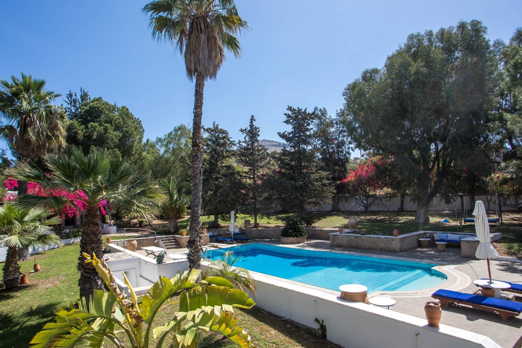 A bright blue swimming pool surrounded by lush greenery and palm trees under a clear blue sky. Lounge chairs are visible. [AI-generated text]