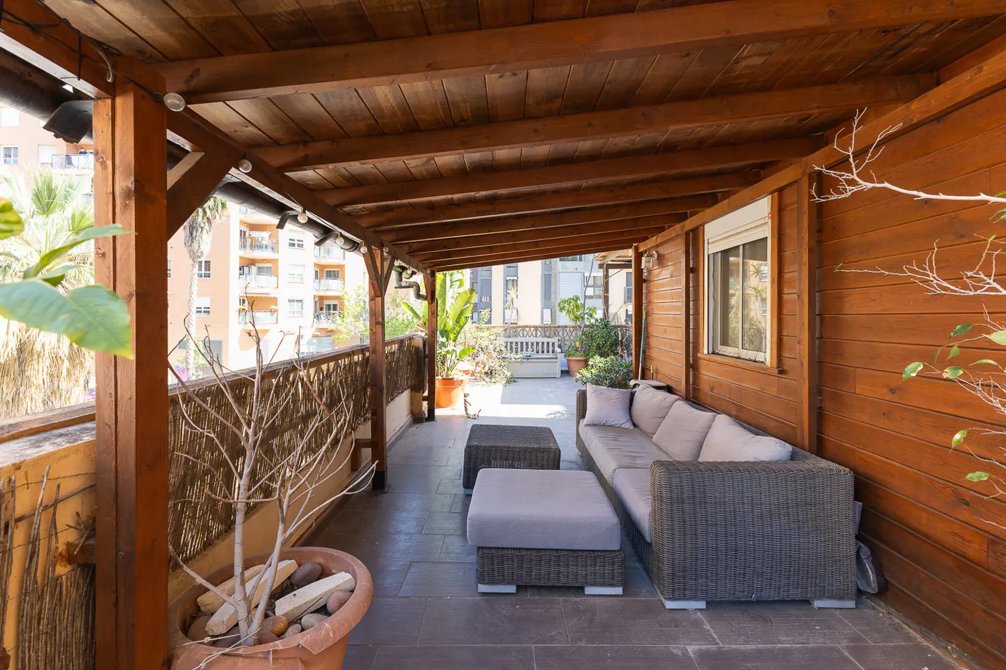 A covered patio with a gray wicker sofa, two ottomans, and a potted plant. The patio has a wooden ceiling and walls.