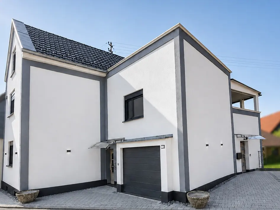 Exterior view of a modern, two-story house with white walls, gray trim, and a dark gray roof and garage door. The house has a small balcony and a paved driveway.