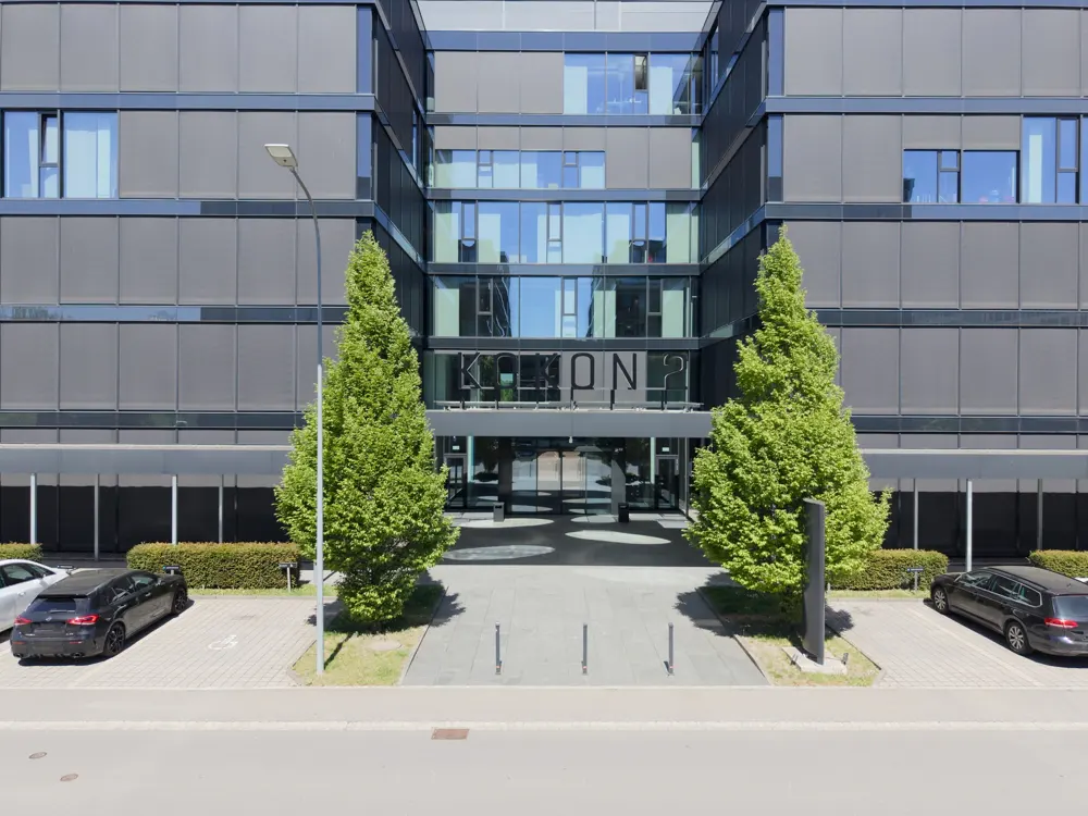 Modern office building with glass windows and gray facade. Two green trees flank the entrance. Cars are parked in the foreground.