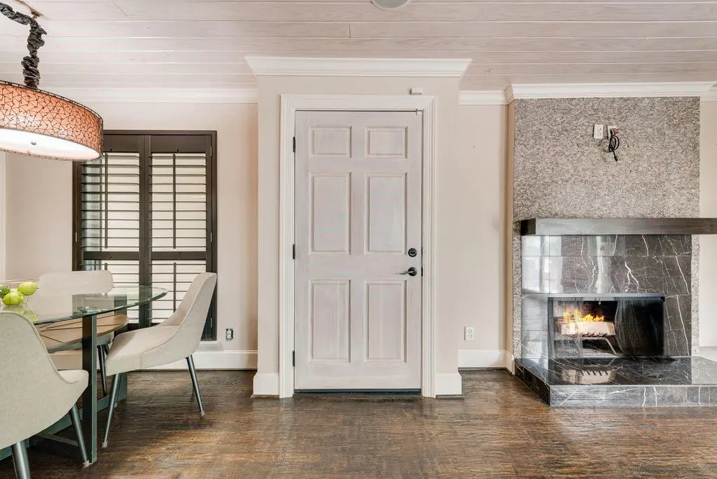 Interior view of a dining area with a glass table, chairs, and a fireplace with a burning fire. A white door is centered.