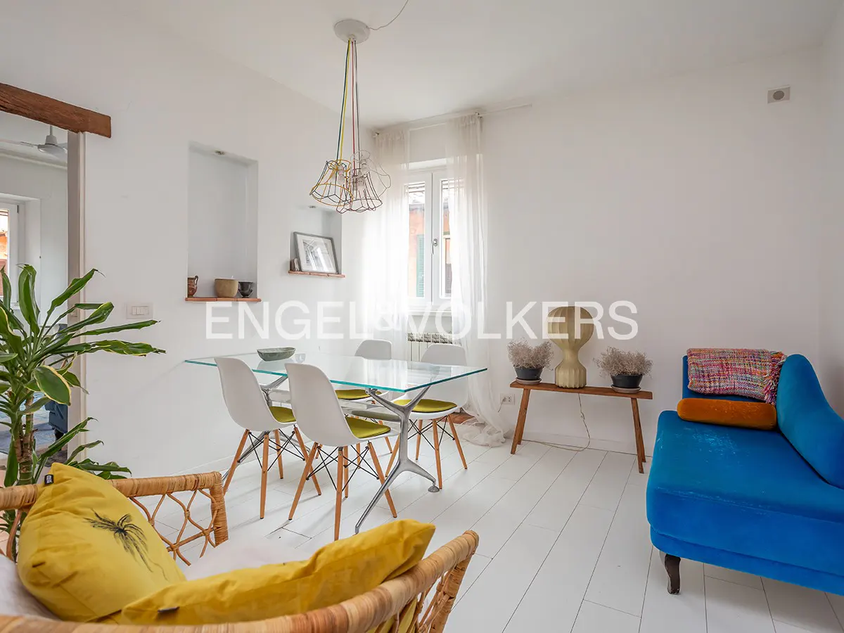 Bright, airy living room with white walls and floors. A glass table with white chairs, a blue chaise lounge, and a wicker sofa with yellow pillows.