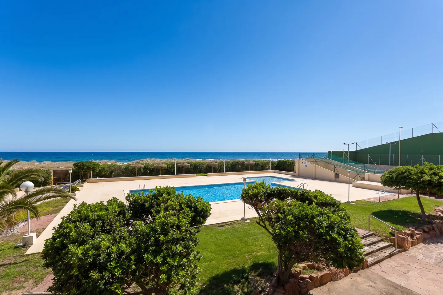 Outdoor pool with blue water, surrounded by green bushes and grass, with the ocean and blue sky in the background.
