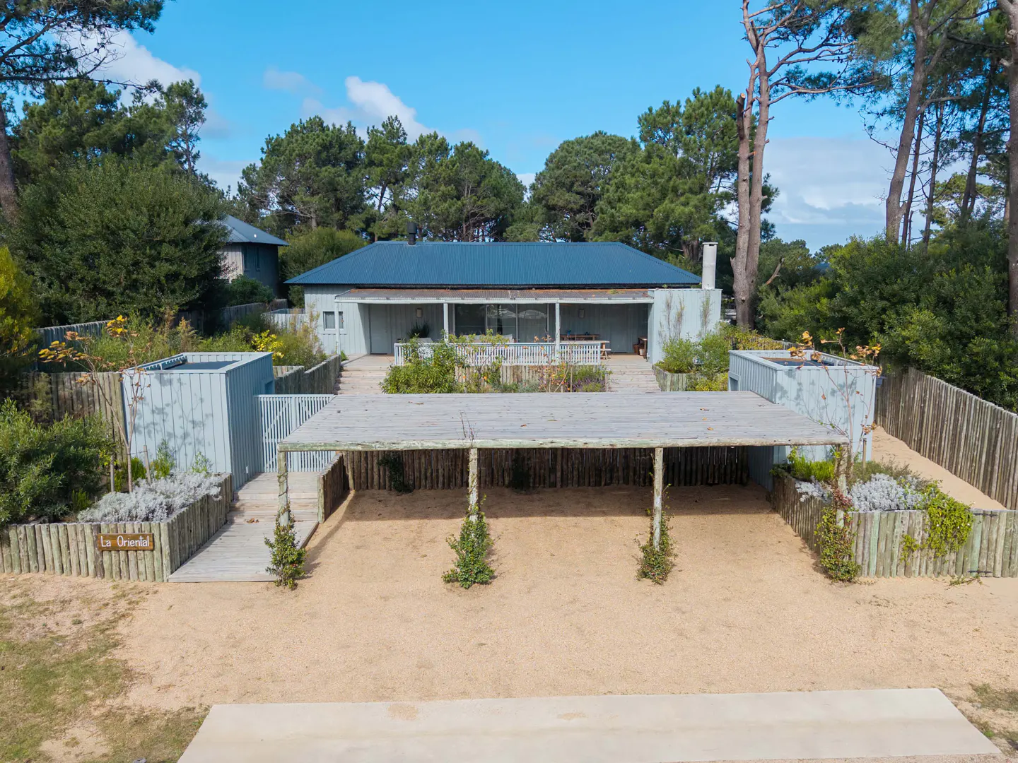 Exterior view of a light blue house with a dark blue roof, surrounded by trees and a sandy yard.