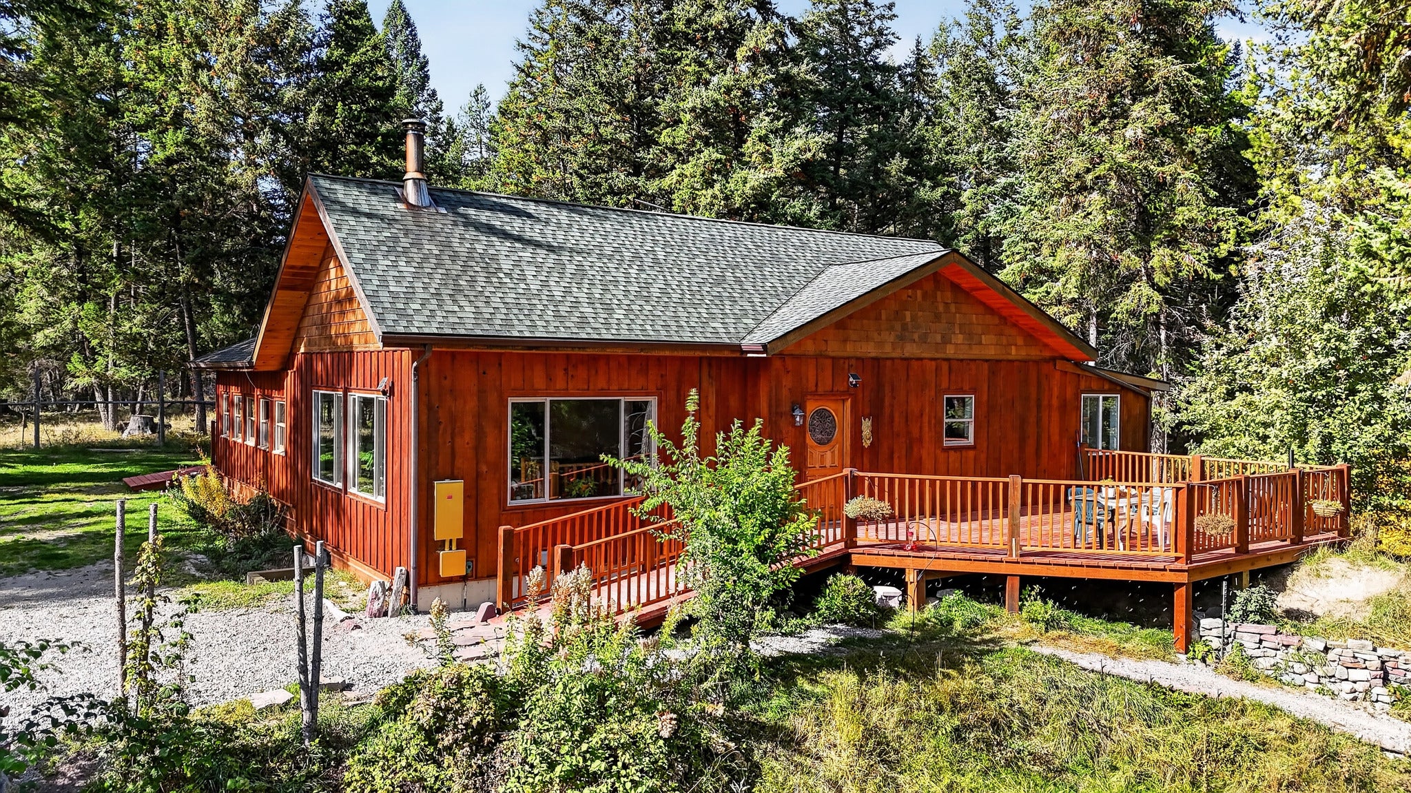 A rustic red wood cabin with a green shingled roof, a wooden deck, and a wheelchair ramp, surrounded by a dense pine forest. [AI-generated text]