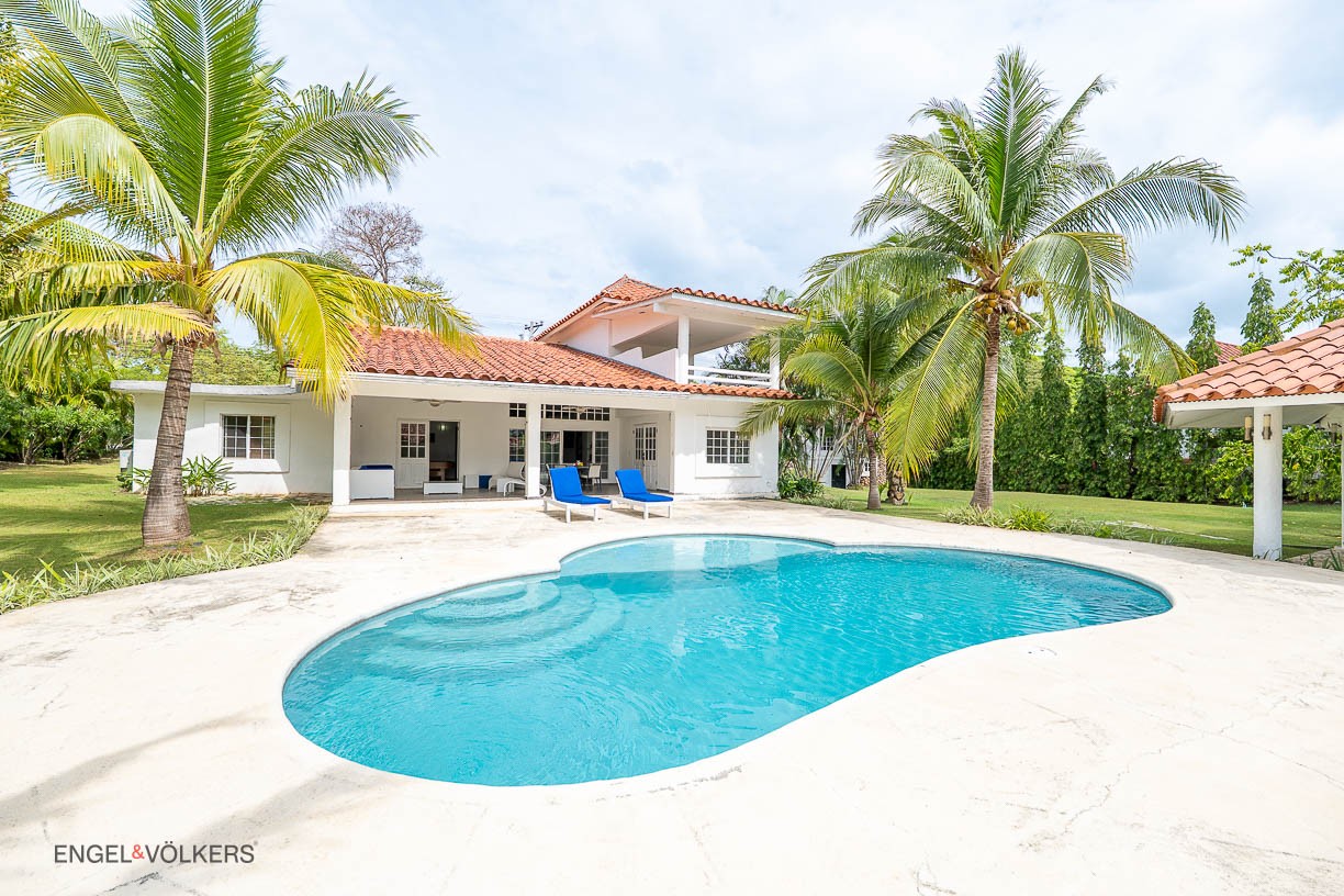 White villa with a red tile roof, turquoise pool, and two blue lounge chairs on a patio surrounded by palm trees. [AI-generated text]