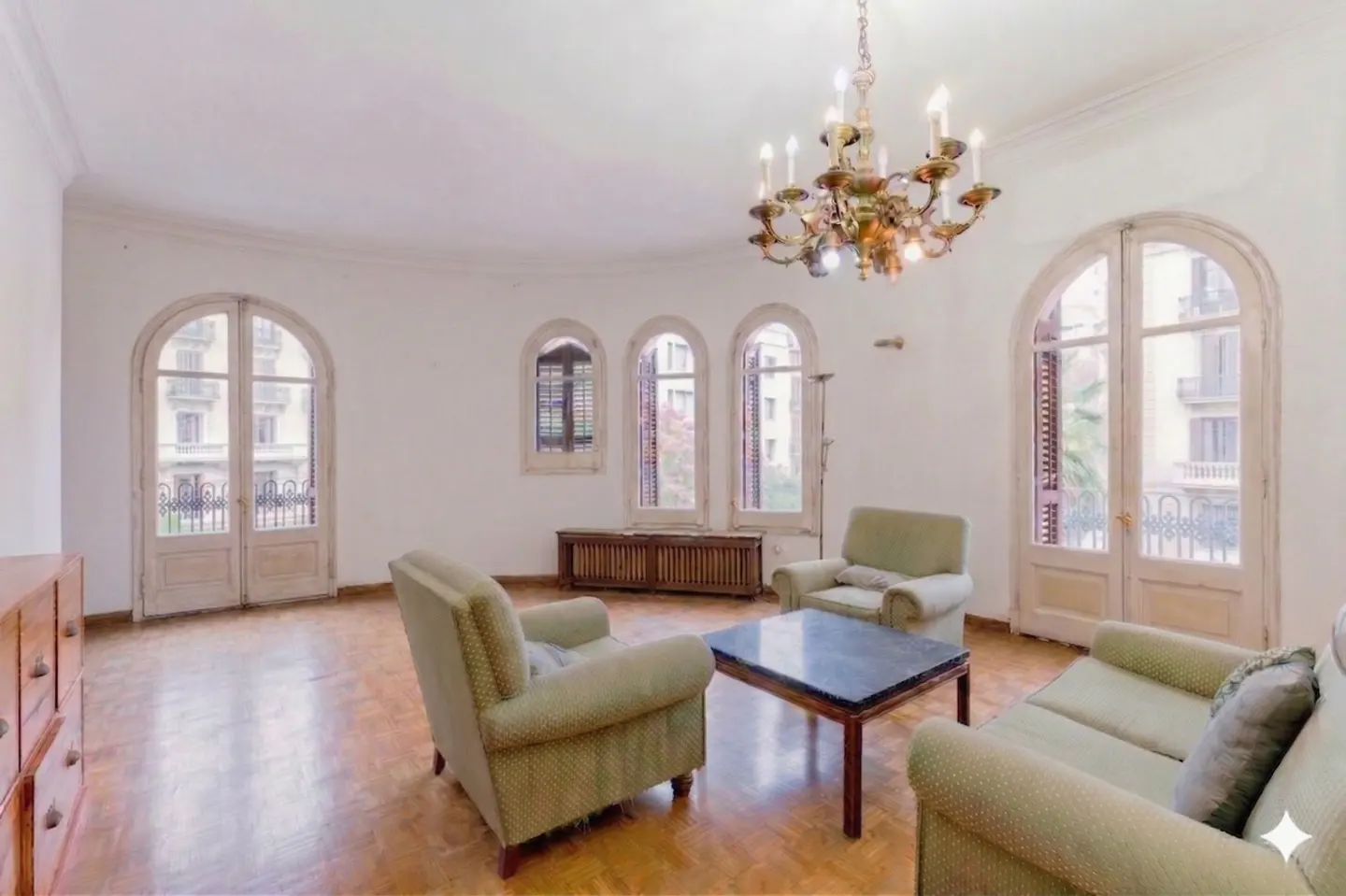 Bright living room with hardwood floors, white walls, and arched windows. A chandelier hangs above green upholstered seating and a dark coffee table.