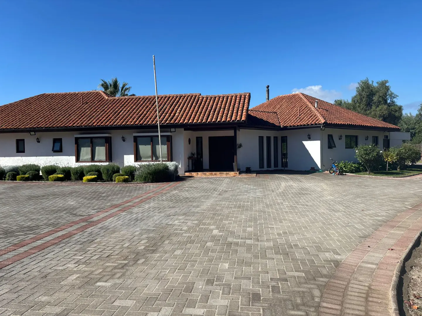 Exterior view of a white, single-story house with a red tile roof and a brick driveway on a sunny day.