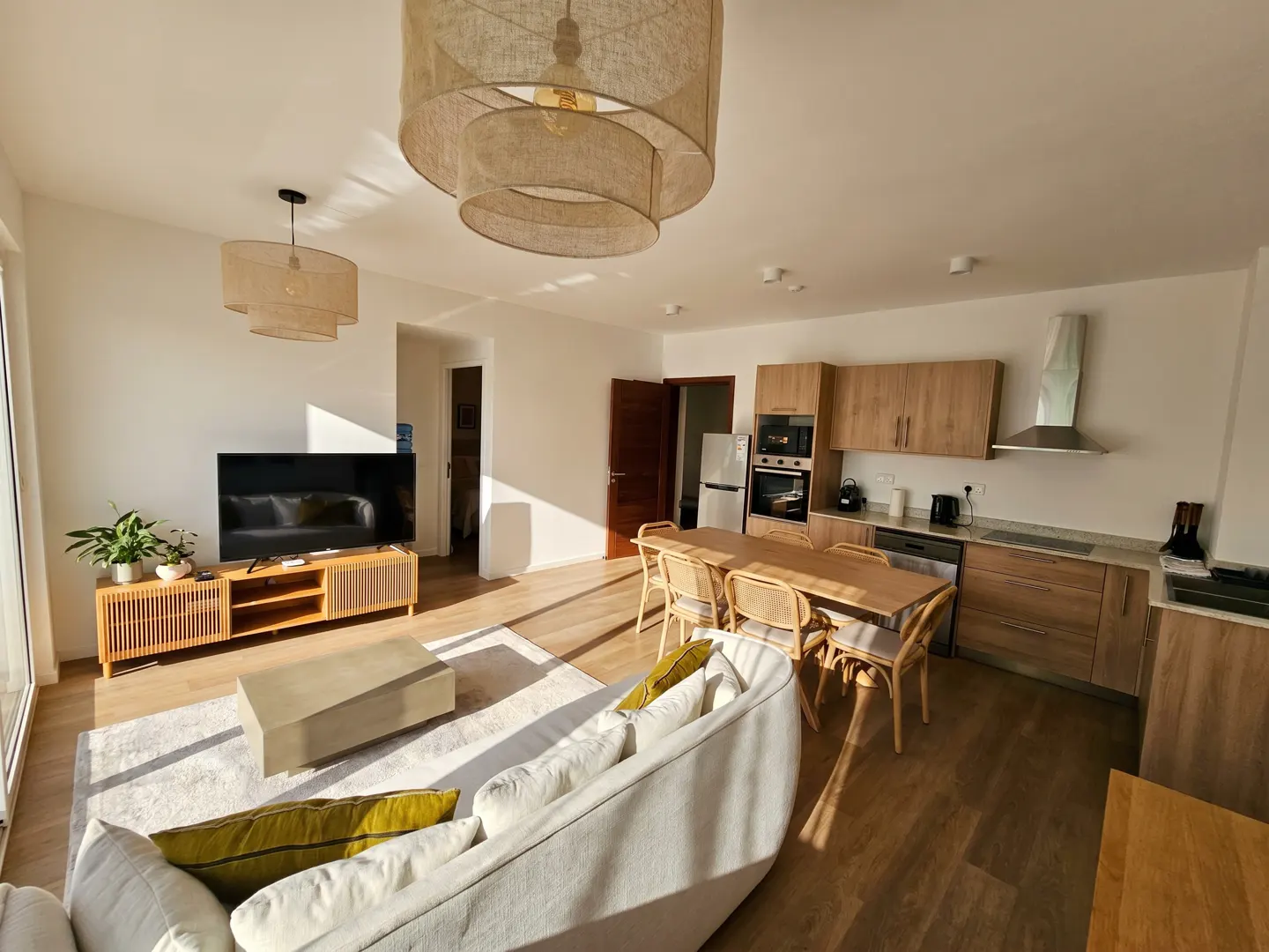 Bright, open-concept living space with a white sofa, wood floors, and a kitchen in the background. Sunlight streams through the windows.