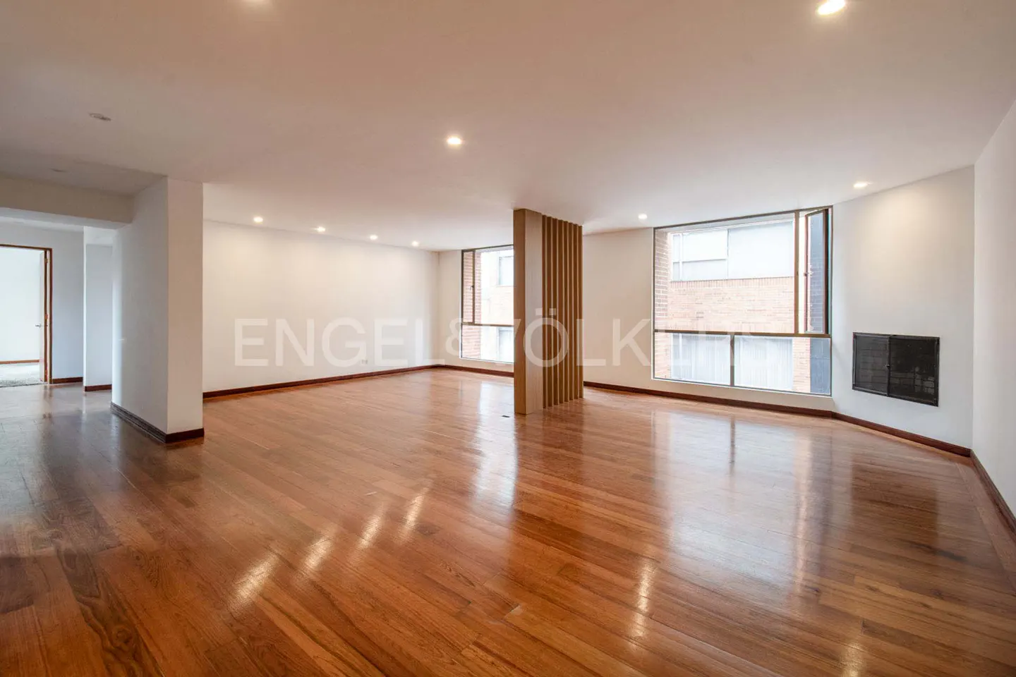 Bright, empty room with hardwood floors, white walls, and large windows. A wooden slat divider stands near the windows.