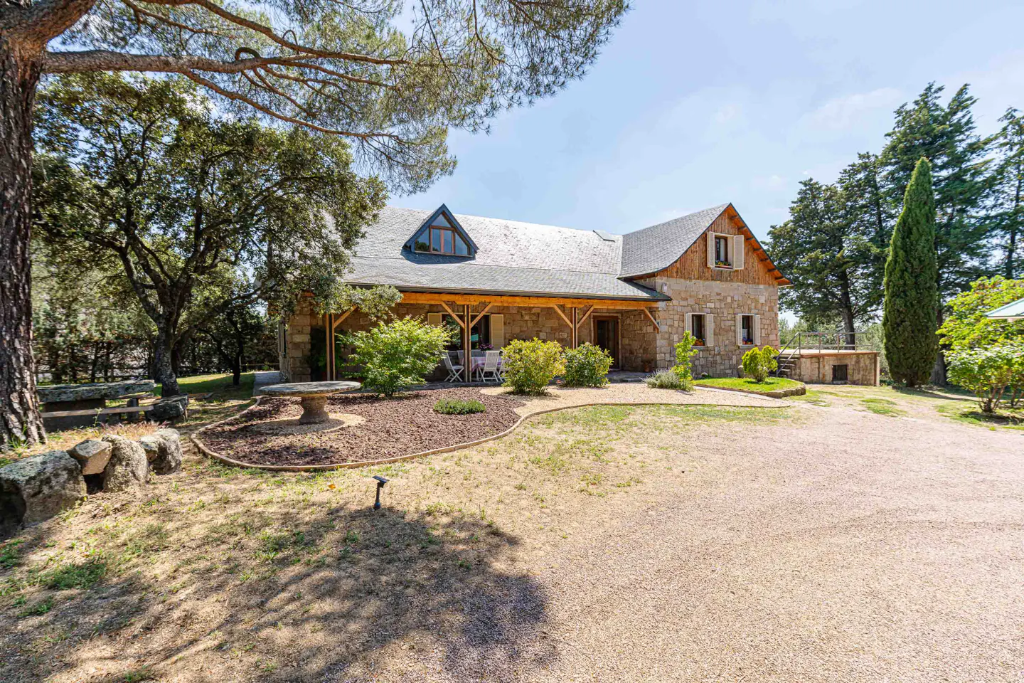 Exterior view of a stone house with a gray roof, wooden porch, and gravel driveway surrounded by trees.