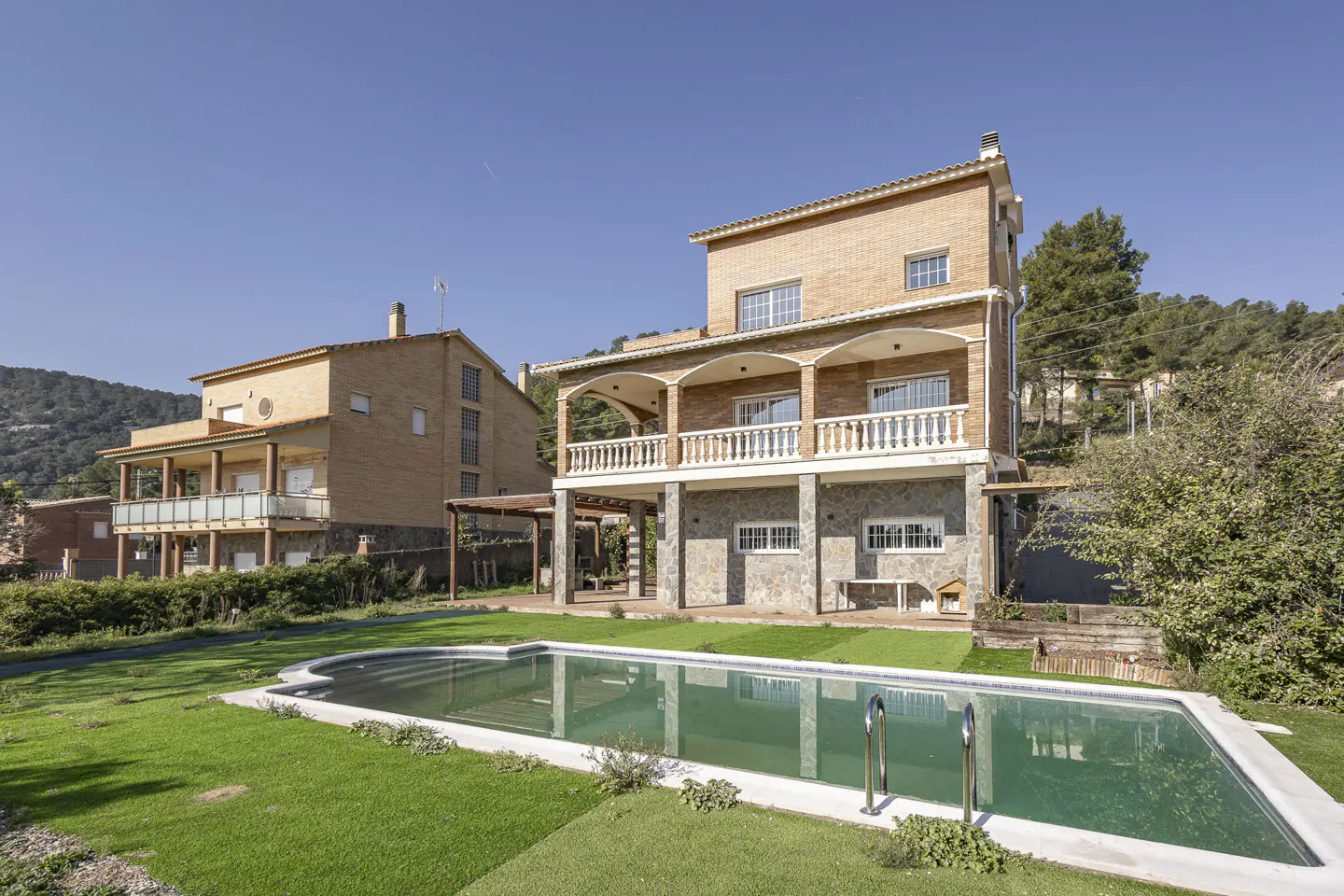 Two-story brick house with a pool. The house has a stone foundation and a white balcony. Green grass surrounds the pool.
