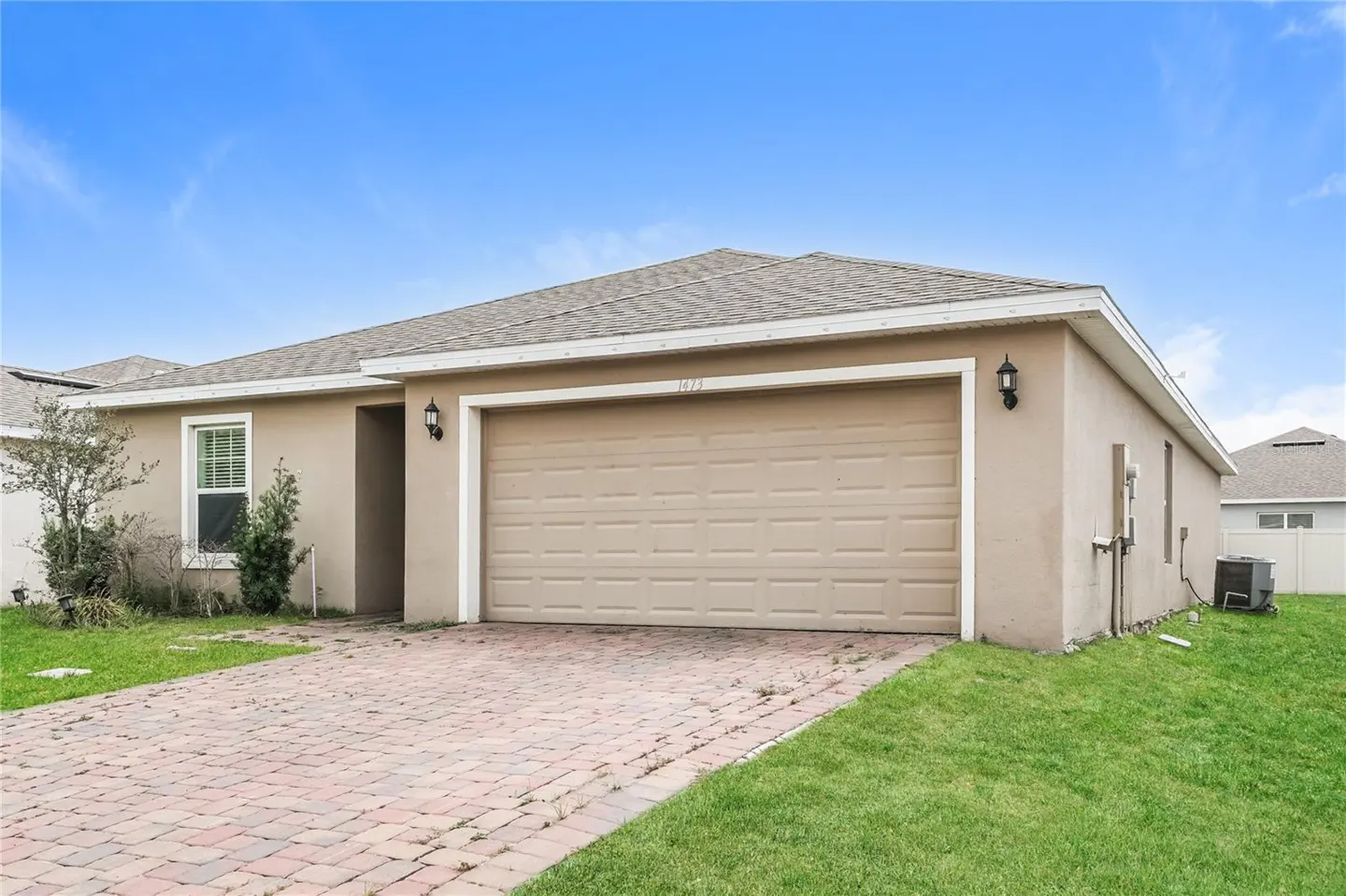 Beige single-story house with a large garage door, brick driveway, and green lawn under a blue sky.