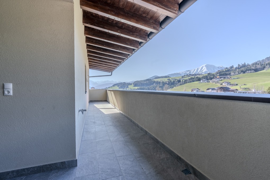 Covered balcony with tiled floor and wooden ceiling beams, overlooking a scenic mountain landscape with green hills. [AI-generated text]