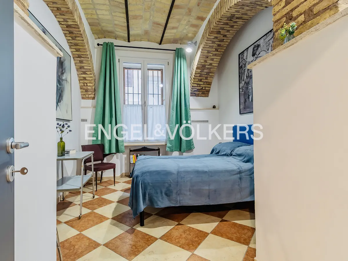 Bedroom with blue bed, green curtains, and checkered floor. Arched brick ceiling and white walls. Doorway on the left.