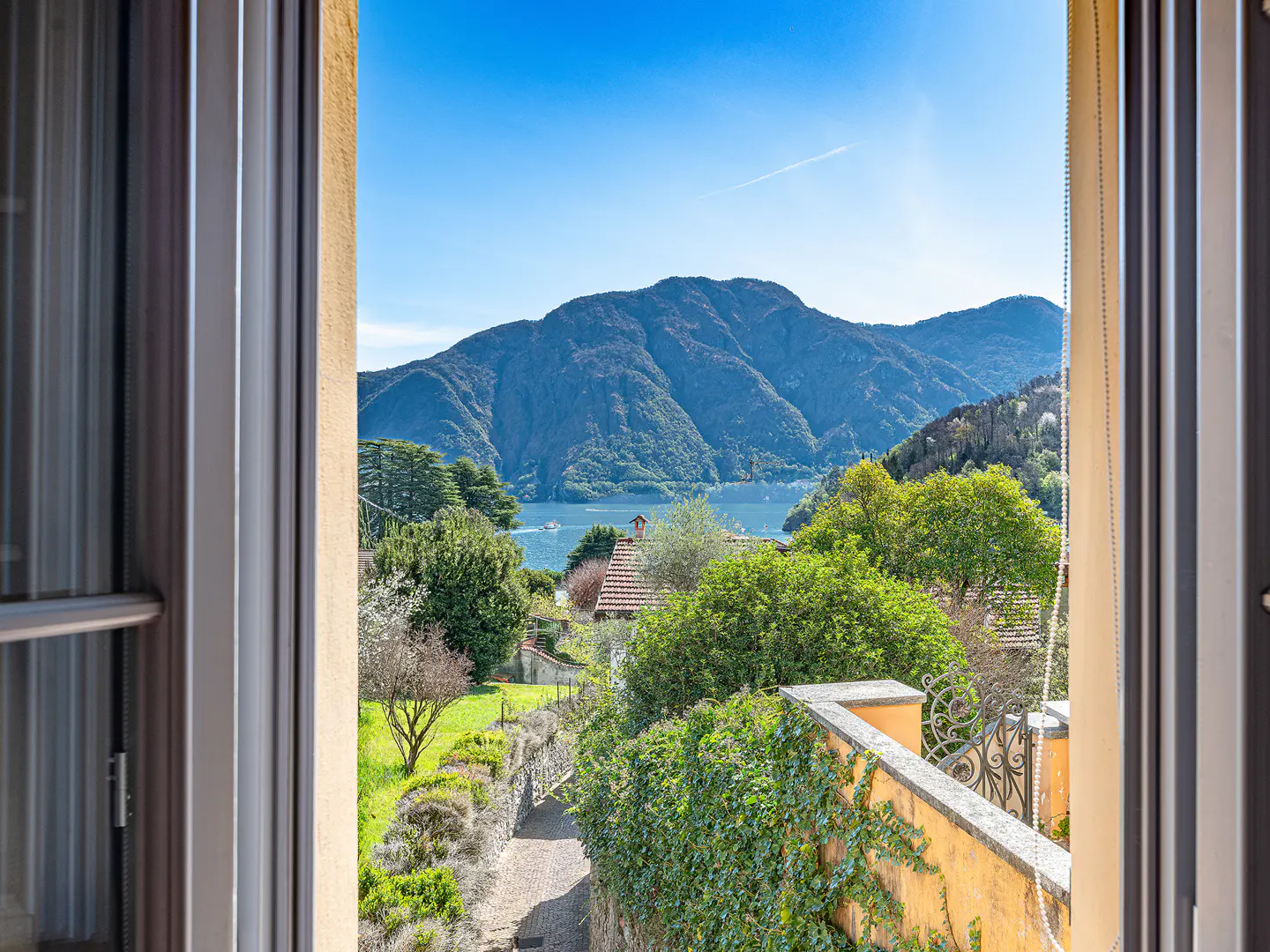 View from a window overlooking a lush green landscape with mountains and a lake under a clear blue sky.