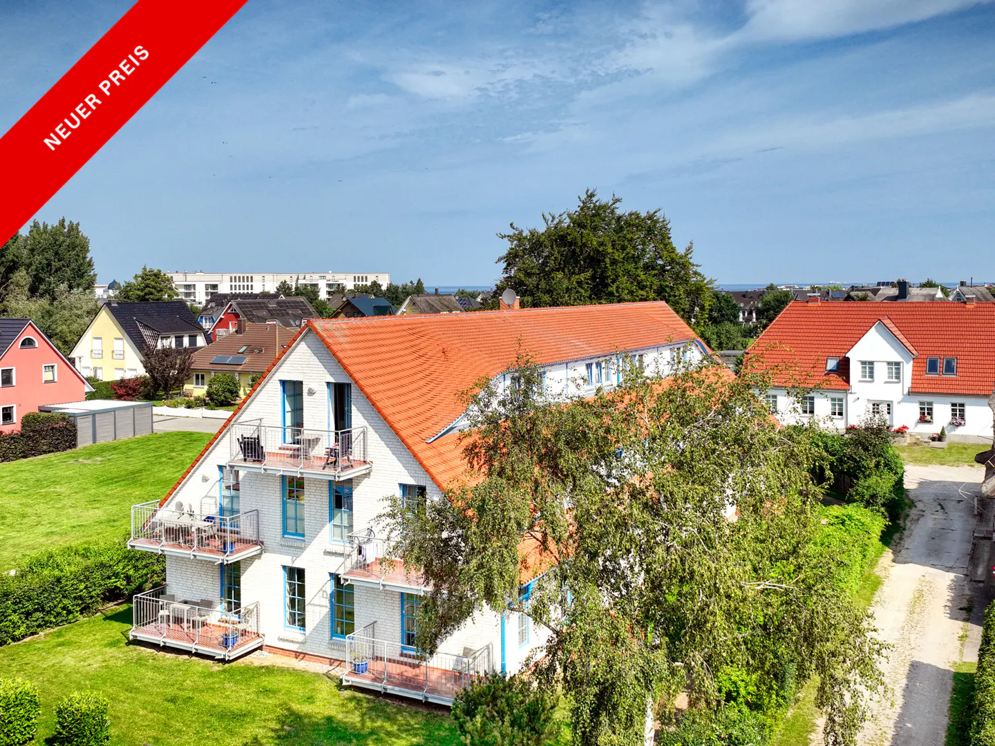 A white building with blue trim and orange roof, surrounded by green grass and trees. "Neuer Preis" banner in the upper left corner.