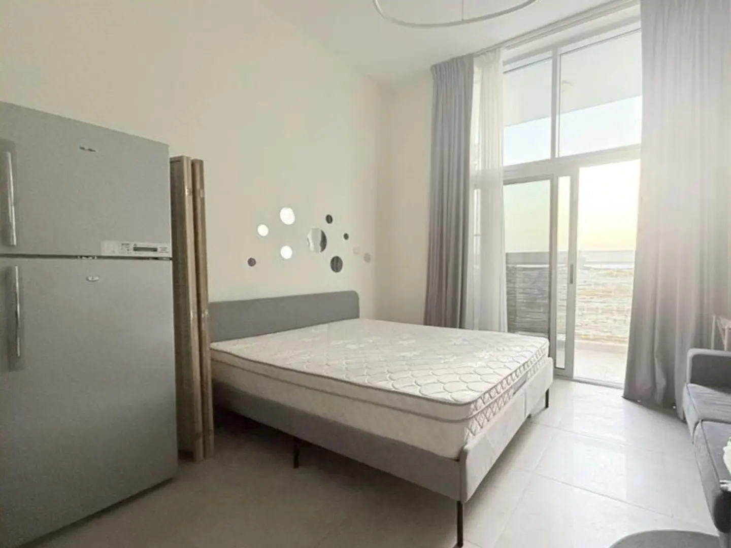 A bright studio apartment with a bed, fridge, and balcony overlooking the ocean. The walls are white, and the curtains are gray.
