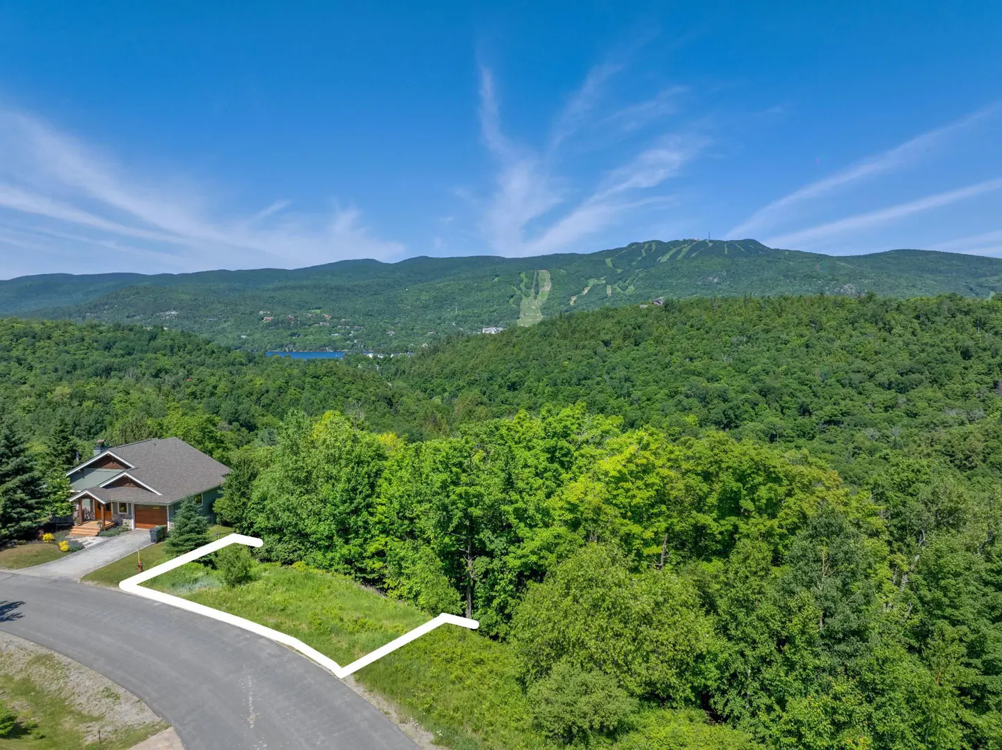 Real estate image showing a vacant lot outlined in white, with a house, road, trees, and mountain views under a blue sky.