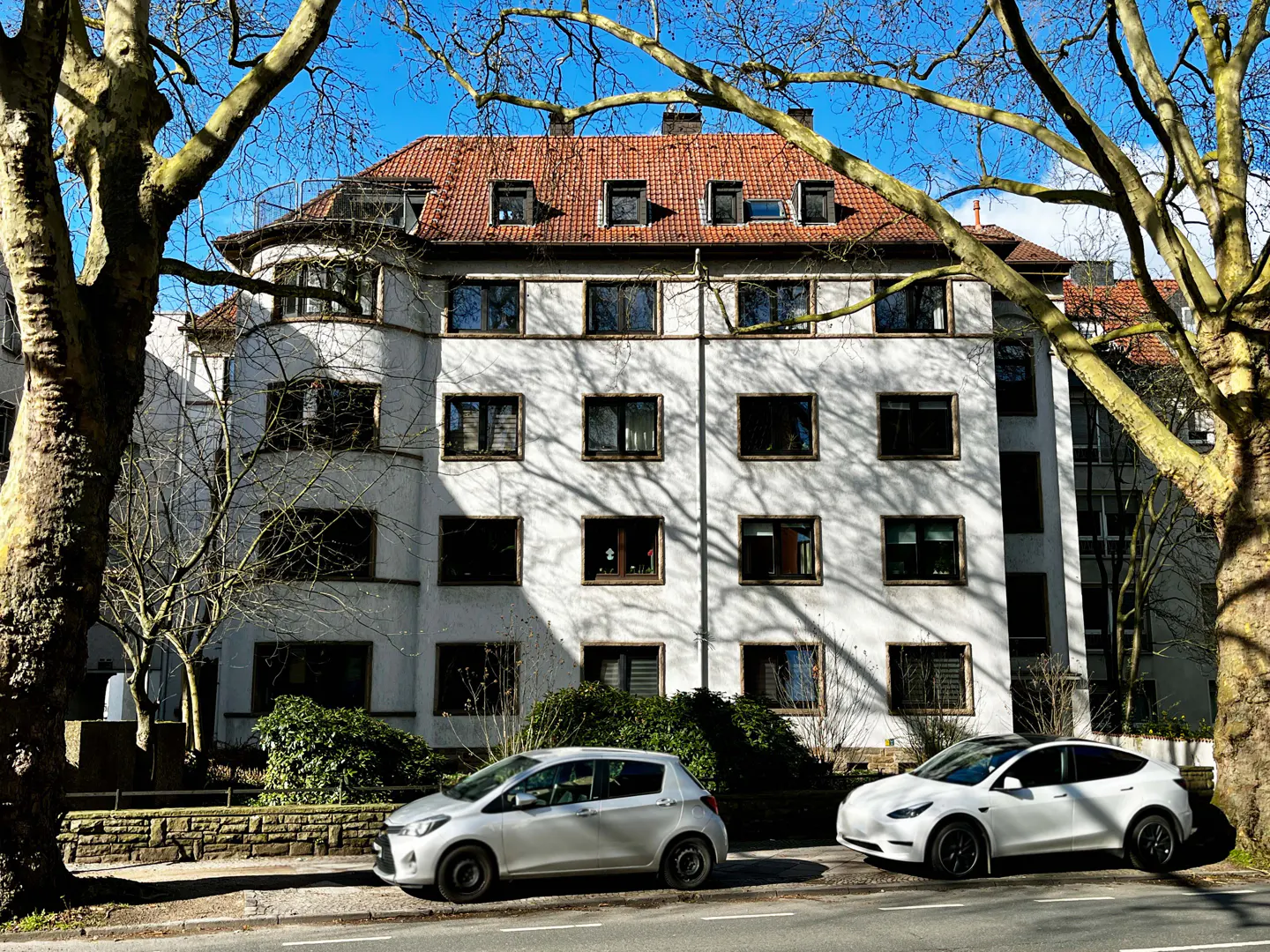 A white apartment building with a red tile roof, framed by bare trees, sits above two parked cars.