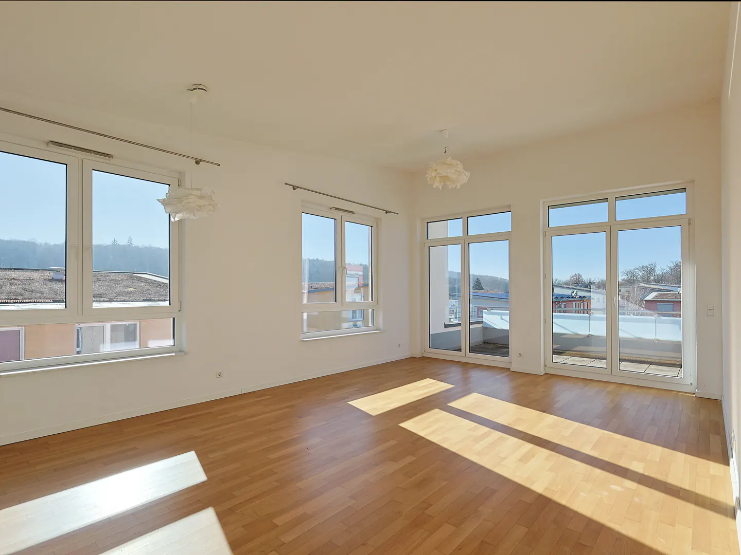 Bright, empty room with wood floors, white walls, and large windows. Sunlight streams in, offering views of trees and buildings. Two white pendant lights hang from the ceiling.