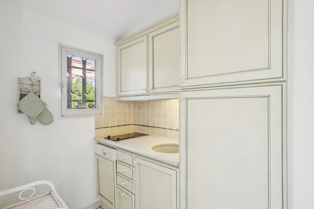 A small kitchen with white cabinets, a sink, and a window. Oven mitts hang on the wall.
