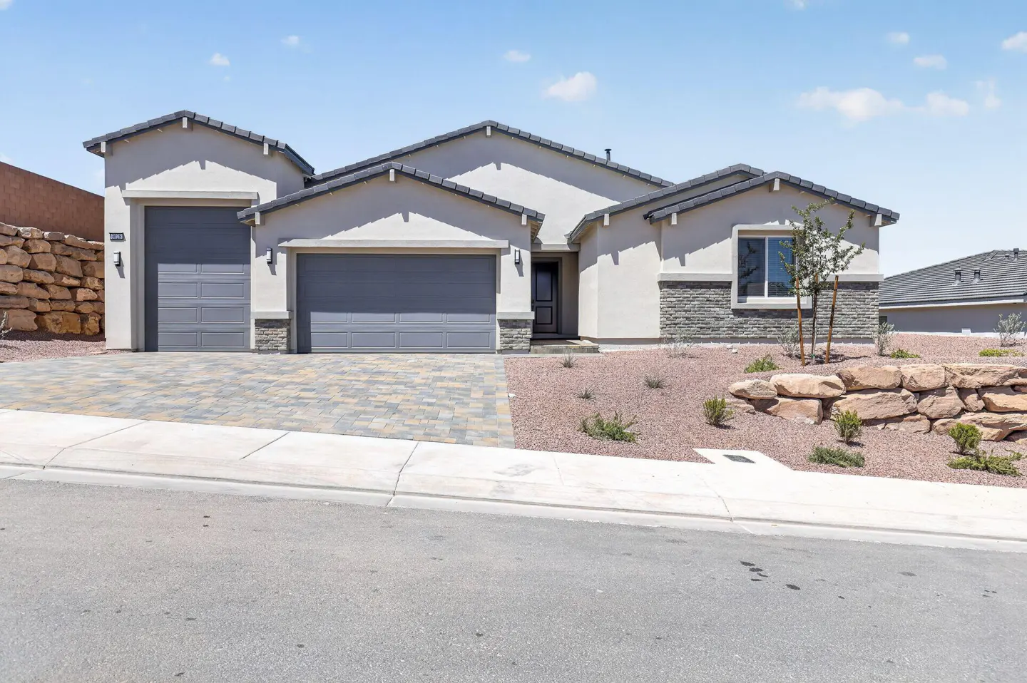 Beige single-story house with gray roof, two-car garage, and stone accents. Landscaped front yard with rock walls and gravel. Blue sky.