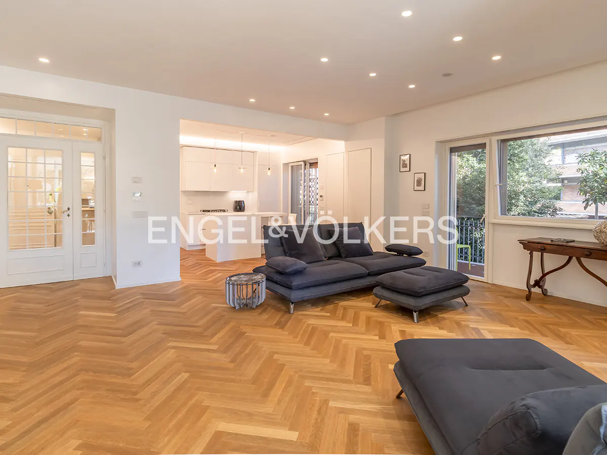 Bright living room with herringbone wood floors, a gray sectional sofa, and a view into a white kitchen. Balcony access and natural light.