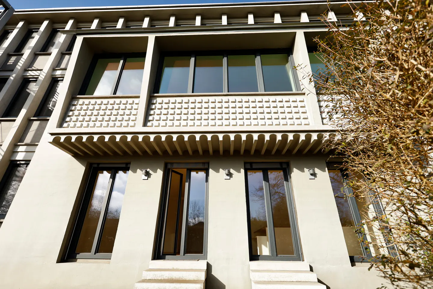 Exterior view of a modern, light gray building with large windows and a decorative balcony. Two sets of steps lead to glass doors. A bare tree is on the right.