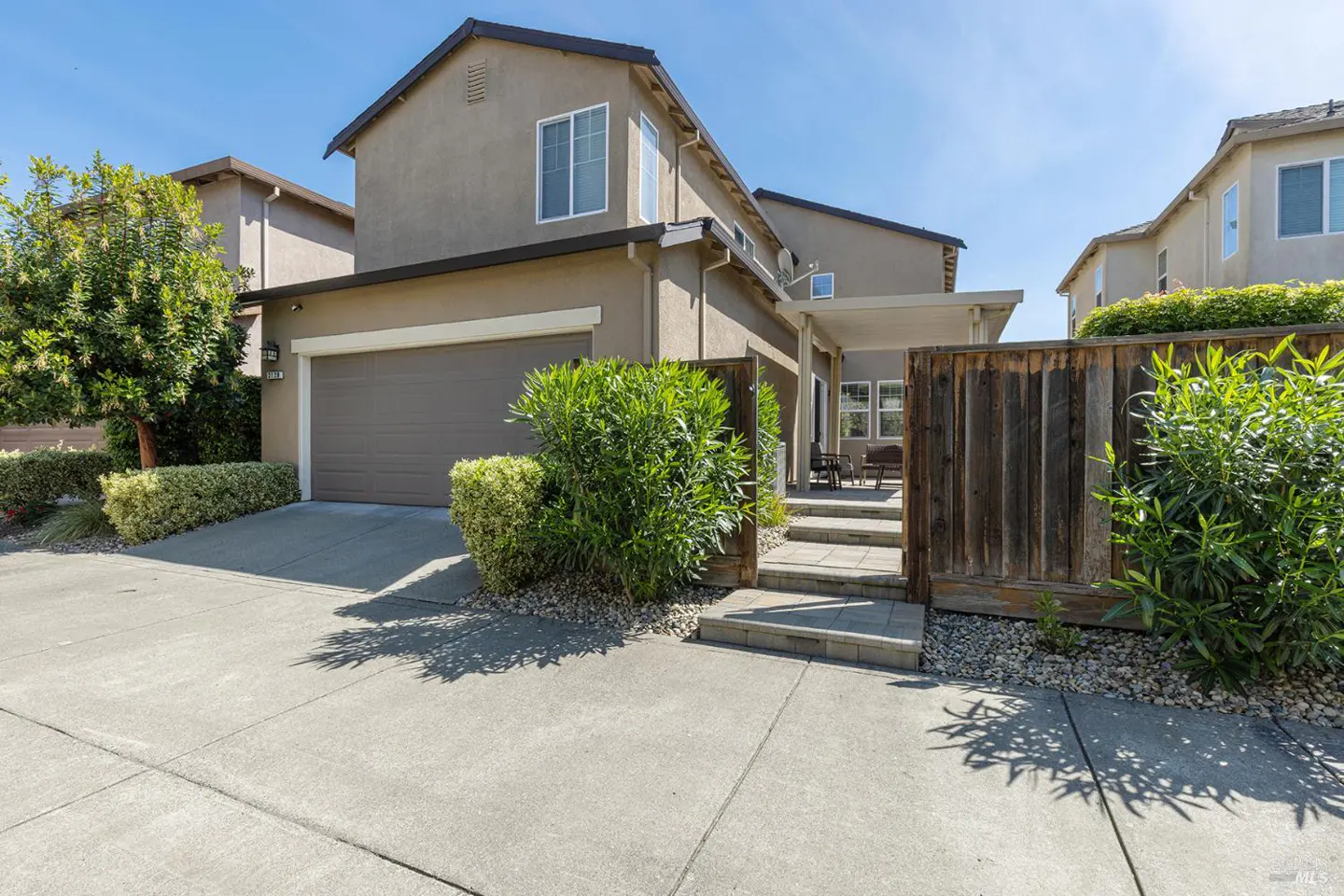 Tan two-story house with a brown garage door, green bushes, and a wooden fence on a sunny day.