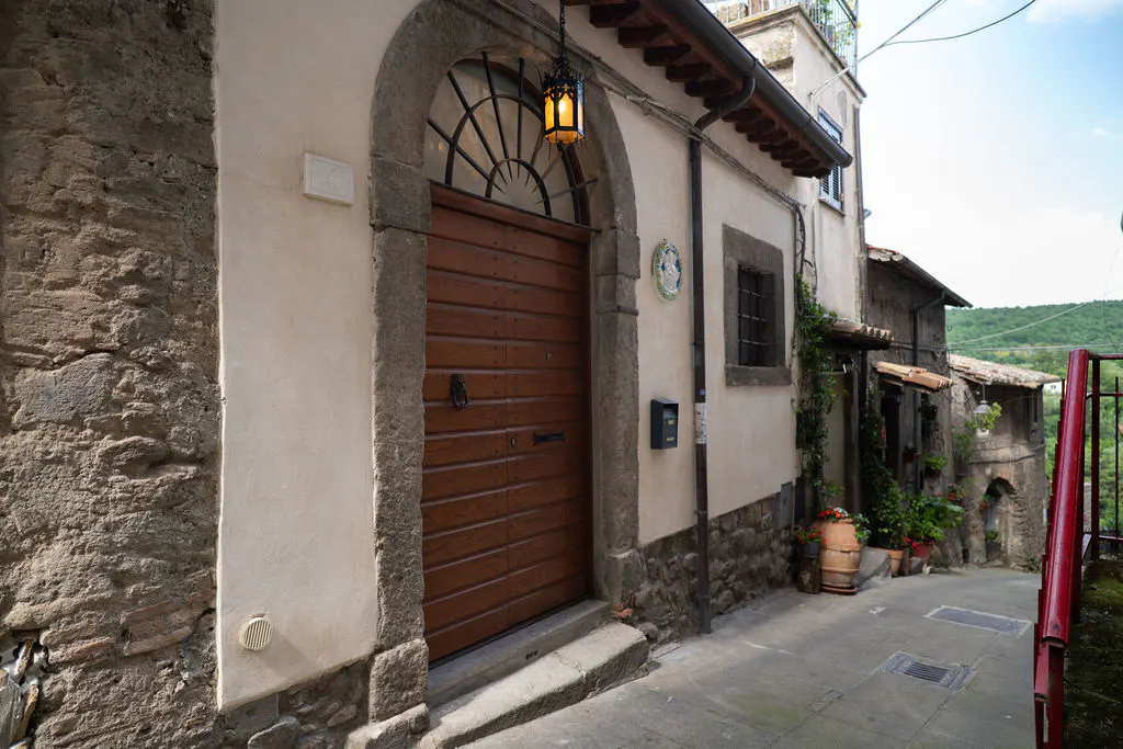 A rustic stone building with a large wooden arched door and a glowing lantern on a narrow, historic Italian village street.
